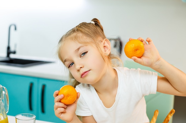 Cute little playful funny girl happily holding orange oranges, with kitchen in the background