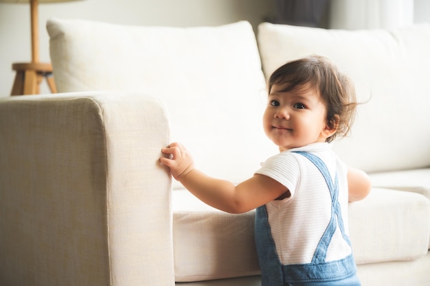 Photo cute little playful girl child learning to walk and stand by taking support of couch at home while looking away mischievously