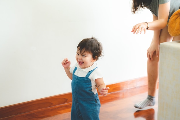 Cute little playful girl child learning to walk and stand by taking support of couch at home while looking away mischievously