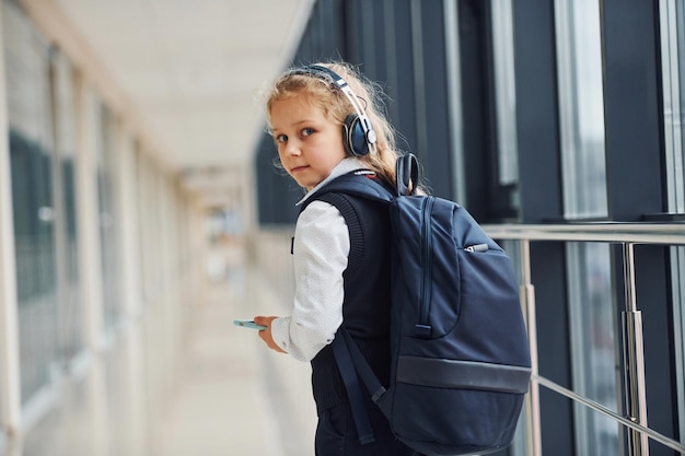 Piccola ragazza sveglia della scuola in uniforme con le cuffie ed il telefono all'interno