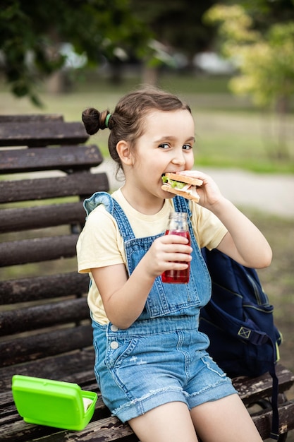 Photo cute little schoolgirl eating from lunch box outdoor sbtting on a bench food for kids