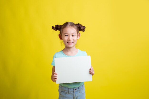 Cute little schoolgirl girl holding a white sheet of paper on a yellow background