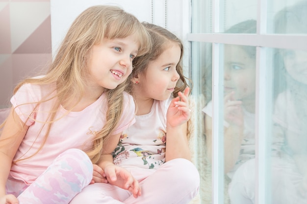 Cute little siblings sitting on windowsill at home looks out window to snow, portrait of pretty little girls on room sill in winter. Adorable pensive child in pink, cute toddler during contemplation.