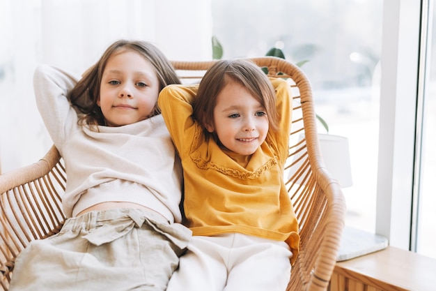 Cute little sisters girls on a wicker chair near window at the home