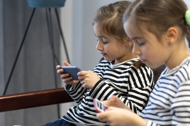 Cute little sisters use phones while sitting on the couch at home.