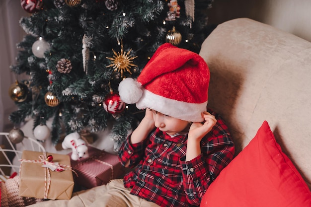 Cute little smiling child kid boy in red hat waiting santa claus in living room with christmas tree