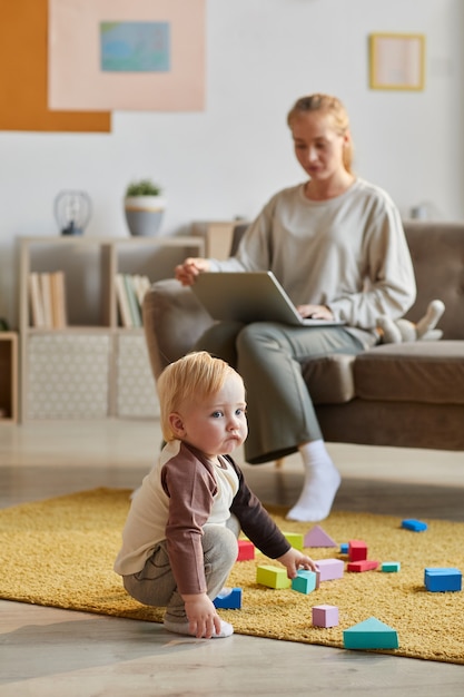 Piccolo figlio sveglio che gioca con i giocattoli sul pavimento mentre sua madre lavora al computer portatile seduto sul divano