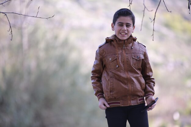 Photo cute little young boy taking selfie using mobile phone camera in the outdoor forest park