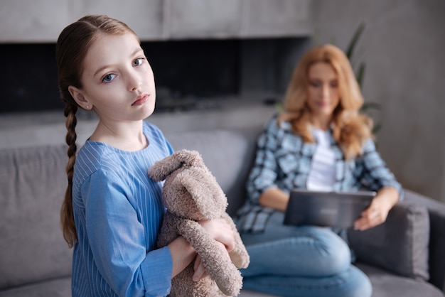 Cute lonely disappointed girl waiting for parent love at home and hugging fluffy toy while mother surfing Internet