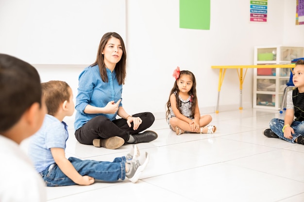 サークルを作って教室の床に座って幼稚園の生徒を教えているかわいらしい若い女性