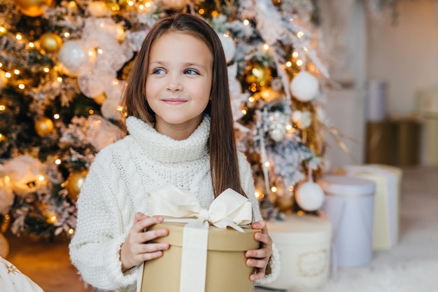 Carino adorabile ragazzino dall'aspetto affascinante felice di ricevere il regalo di natale guarda da parte con un'espressione felice ha intriga ciò che il regalo è in posa vicino al bellissimo abete decorato