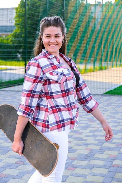 Photo cute millennial woman smile happy walking down the street with her skateboard sun backlight