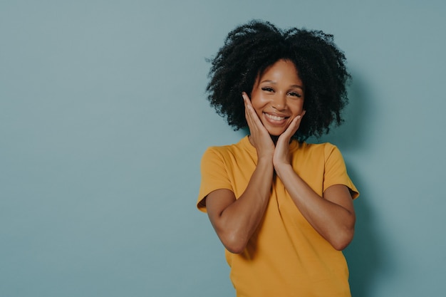 Cute mixed race young woman touching cheeks with hands and feeling happy after hearing compliment