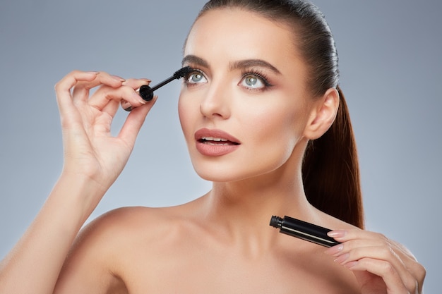 Cute model with brown hair fixed behind, clean fresh skin, big eyes and naked shoulders posing at gray studio background and looking at camera, close up, holding mascara.