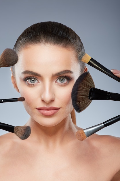 Cute model with brown hair fixed behind, clean fresh skin, big eyes and naked shoulders posing at gray studio background and looking at camera, close up, using make up brushes.