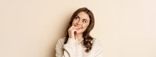 Cute modern woman thinking daydreaming and smiling looking up\
thoughtful standing over beige backgro