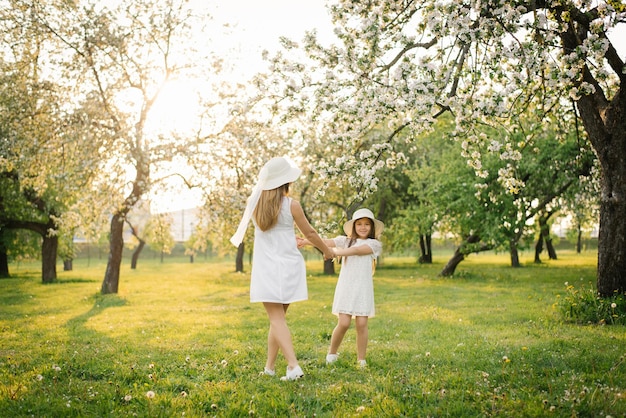 A cute mom in a hat and a white dress is walking through the spring garden with her cute daughter