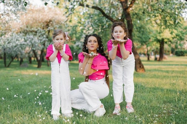 Милая мама и две дочери дуют на опавшие лепестки яблоневых цветов в весеннем саду