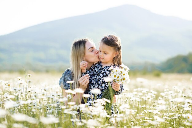 Cute mother kissing child girl daughter in meadow with blooming flowers outdoor together