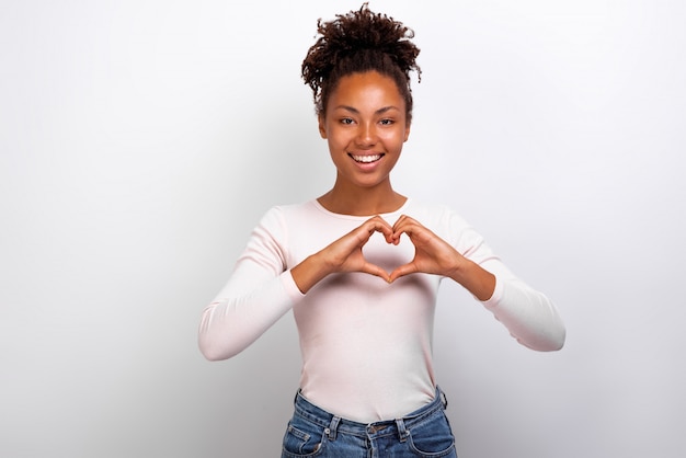 Cute mulatto woman shows a heart gesture with her fingers next her chest. 