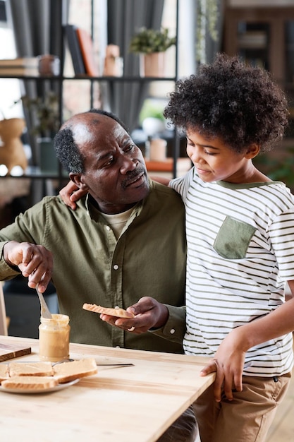 Cute multiethnic schoolboy standing by his grandfather making him sandwich