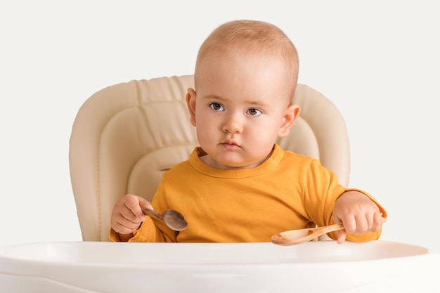 A cute newborn baby aged one year is sitting on a feeding chair in orange casual clothes and holding two wooden spoons in his hands while waiting for lunch.