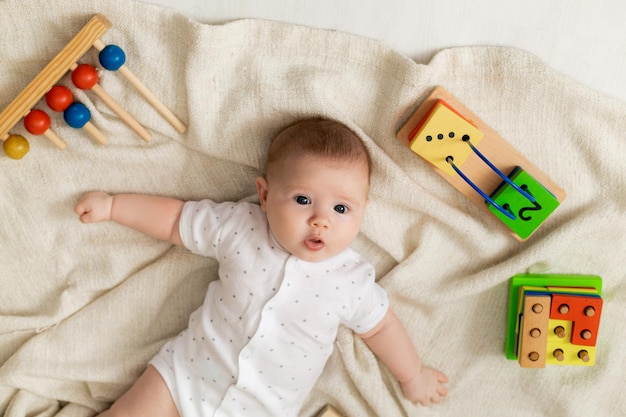 薄着のかわいい新生児は、灰色のリネンの毛布の上に床に横たわっている知育おもちゃで遊んでいます。上面図。子供のための製品。幸せな子供時代と母性。テキスト用のスペース