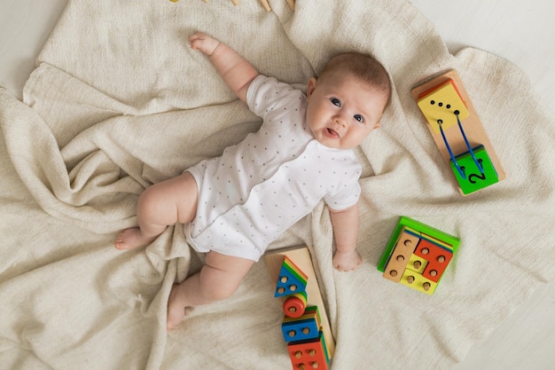 Cute newborn baby in light clothes is playing with educational toys lying on the floor on a gray linen blanket. top view. products for children. happy childhood and motherhood. space for text