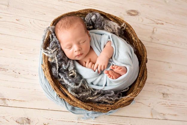 A cute newborn boy in the first days of life sleeps in a blue winding wicker brown basket on a light wood background high quality photo