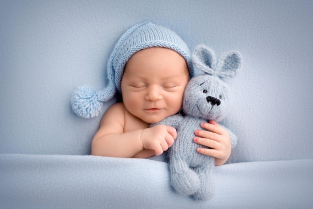 A cute newborn boy in the first days of life sleeps naked on a blue fabric background The kid gently hugs a light blue knitted bunny Studio professional macro photography newborn baby portrait