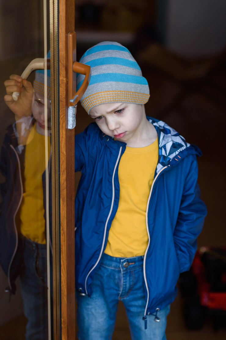 Cute offended boy stands on the doorstep and holds on to the door handle