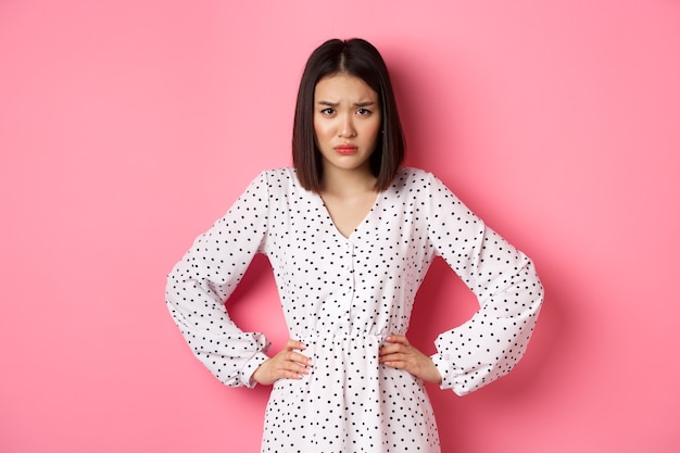 Cute offended girl frowning, holding hands on waist and staring at camera disappointed, standing upset and gloomy over pink background.