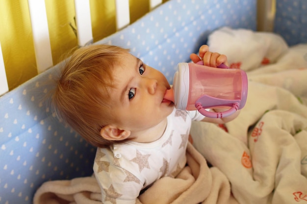 可愛い1歳の女の子が朝起きてベッドでボトルから水を飲んでいます