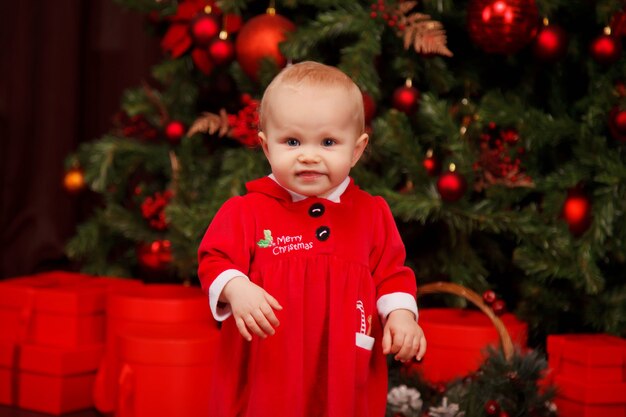 크리스마스 트리에 산타 클로스 의상을 입은 귀여운 한 살짜리 소녀가 장난감으로 장식되어 있습니다. 크리스마스 훈장에있는 아이