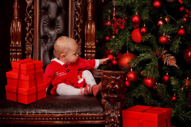 Cute one-year-old girl in Santa Claus dress on throne on background of Christmas tree decorated toys. Child in decorations with gift boxes. Concept of cozy home Happy New Year celebration. Copy space