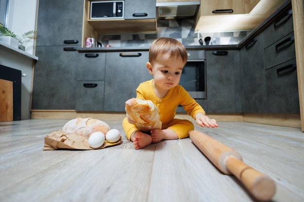 A cute oneyearold boy is sitting in the kitchen and eating a long bread or baguette in the kitchen The first eating of bread by a child Bread is good for children