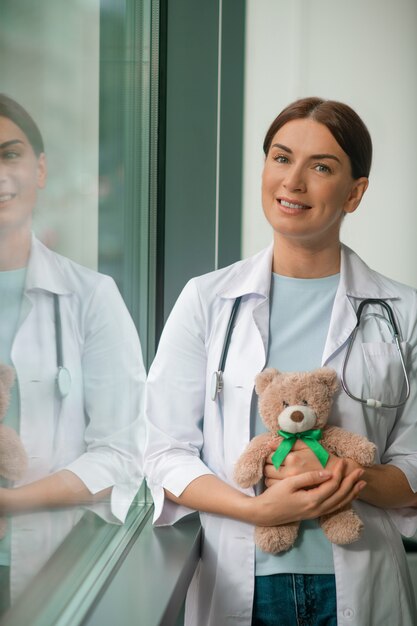 A cute pediatrician standing near the window and holding a teddy bear