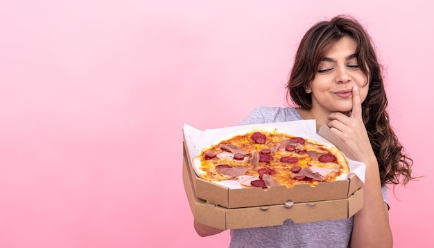 Cute pensive girl with a box of pizza for delivery on a pink background, copy space.