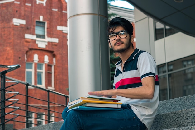 Ragazzo carino e pensieroso con gli occhiali che tiene un libro e guarda avanti