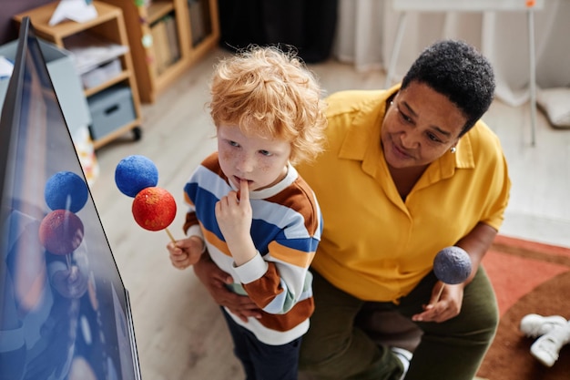 Cute pensive little boy with models of earth and mars looking\
at interactive board at lesson while s