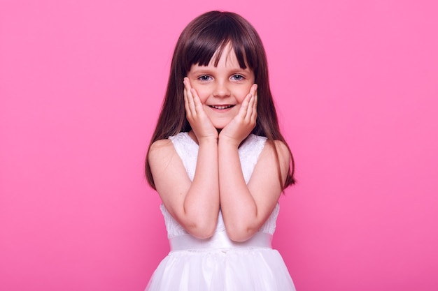 Симпатичная симпатичная маленькая девочка в стильном белом платье, смотрящая вперед с очаровательным счастливым выражением лица, довольная, любуясь чем-то, изолированная над розовой стеной