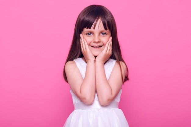 Cute pleasant looking small girl wearing stylish white dress looking at front with charming happy expression, being satisfied, admiring something, isolated over pink wall