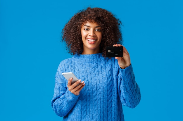 Photo cute pleased african-american female bank customer recommend credit card and banking system service, holding smartphone and smiling, buying online, shopping in internet stores, blue background