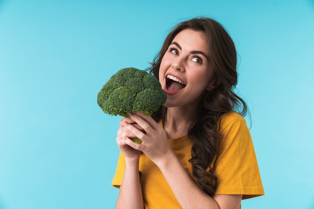 cute pleased young beautiful woman posing isolated over blue wall holding broccoli.