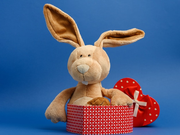 Cute plush rabbit holding a gift box tied with a red silk ribbon, blue background, close up