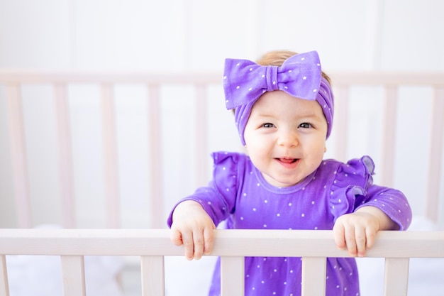 Cute portrait of a baby girl in a crib holding on to the side in lilac clothes and with a bow on her head and laughing the concept of children's goods