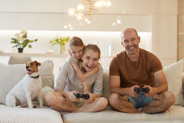 Cute portrait of happy modern family playing video games together while sitting on couch in cozy home interior, smiling mature man enjoying time with two daughters and pet dog