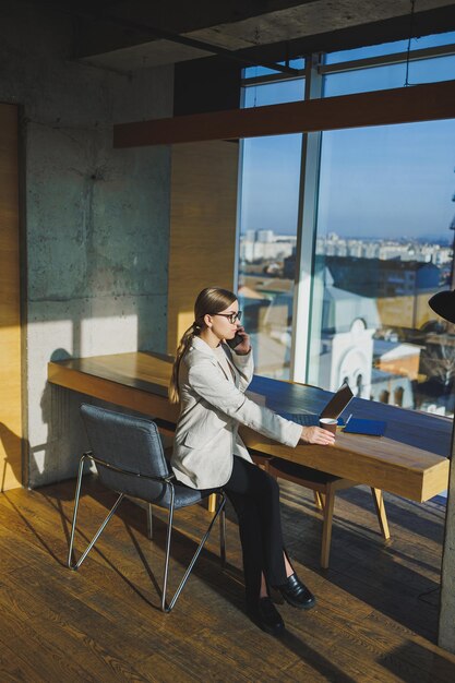 Cute positive female employee with long blond hair in casual clothes looking at phone while working on new business project at table with laptop and gadgets in office