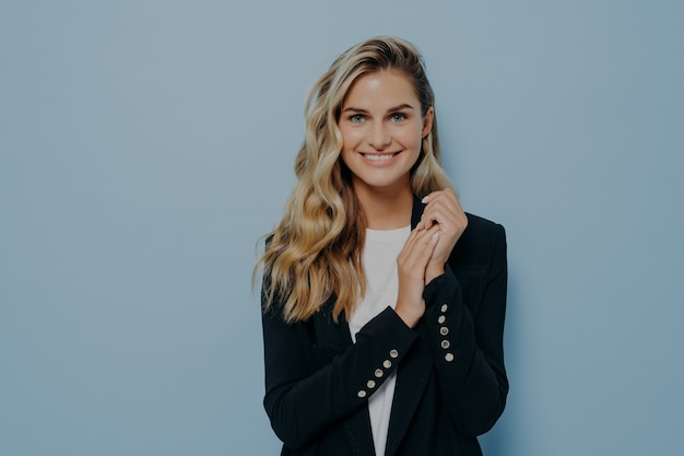 Cute positive female student in stylish casual outfit posing against blue studio wall, looking happily at camera and expressing positivity. Beautiful and happy young people concept