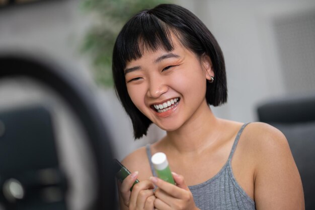 Cute positive video blogger with the skincare products in her hands looking at the smartphone camera